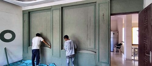 Two workers finishing sage-green wall panelling, with a red door and a fridge visible through the opening beyond.