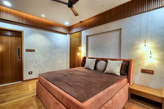 A terracotta bed under a stone headboard panel, with ribbed timber running across the ceiling and two wall lights.