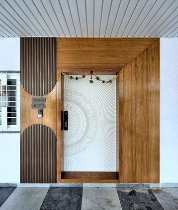 An arched teak front door with a carved white panel set into it, under a slatted timber ceiling.