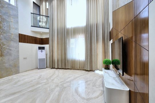 The living room from its far corner — a teak panel carrying the television, a white console below it, tall windows and a marble floor.
