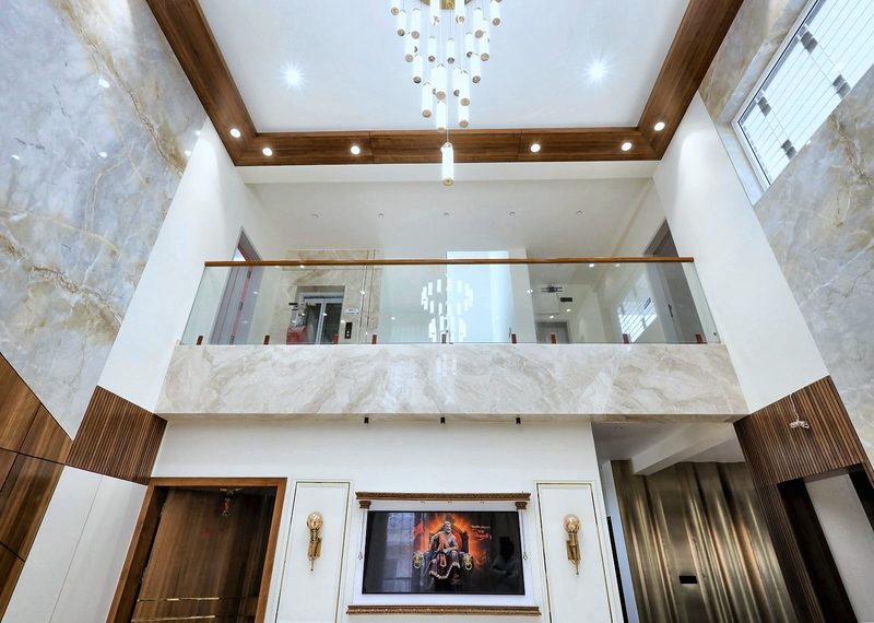 The double-height living room photographed from the first-floor landing — the glass balustrade in the foreground, a chandelier hanging the full height of the void, the stone and teak wall below.