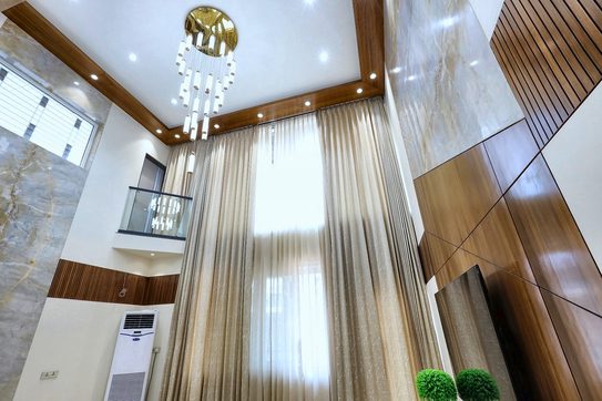 The double-height living room with sheer curtains running the whole height of the wall, a chandelier above and the stone and teak panel to the right.